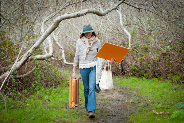 Person walking in a forest holding a painting board and a bag.